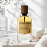 A clear glass perfume bottle filled with pale yellow liquid, featuring a natural wood cap and a tan paper label that reads 'Liquid diamonds'. The bottle is styled on a white marble surface with clear crystals and a soft white fabric in the background.