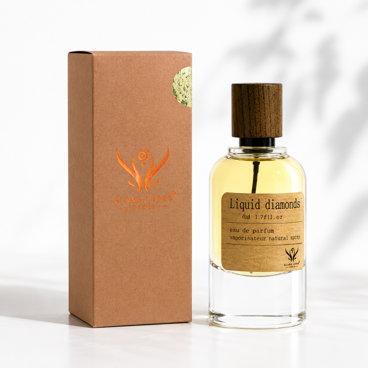 A clear glass perfume bottle filled with pale yellow liquid, featuring a dark wood cap and a tan label reading 'Liquid diamonds', standing next to a brown kraft paper box with an orange logo and a small green seal. The background is a clean white with soft leaf shadows.
