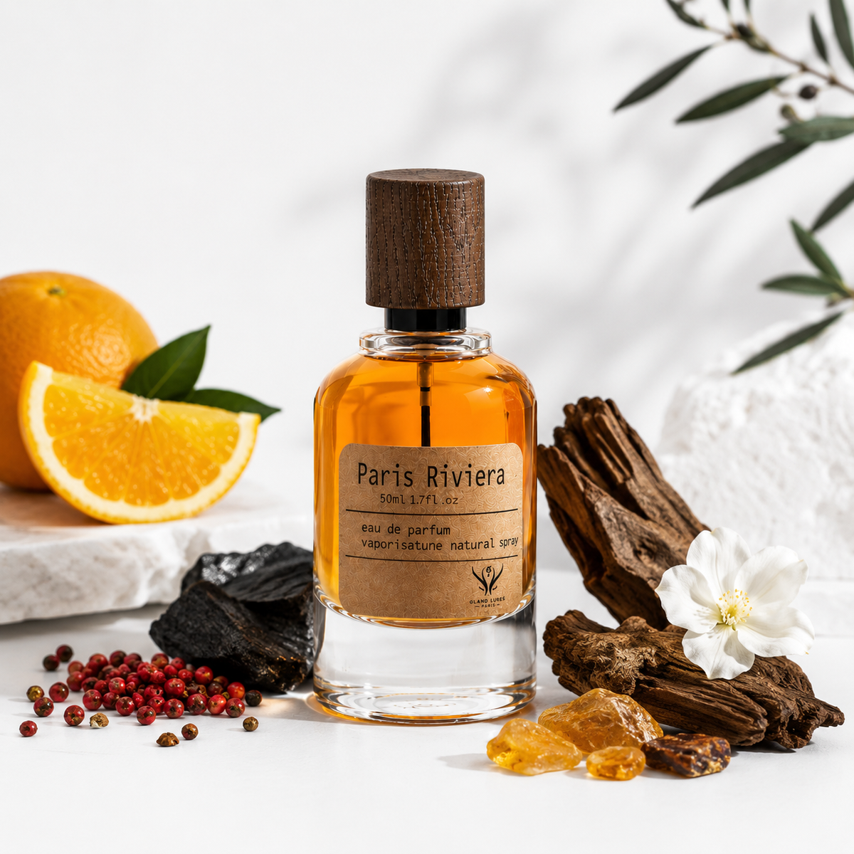 A perfume bottle labeled Paris Riviera, filled with amber liquid, set on a white surface with an orange slice, red peppercorns, dark wood pieces, amber crystals, and a white flower around it.