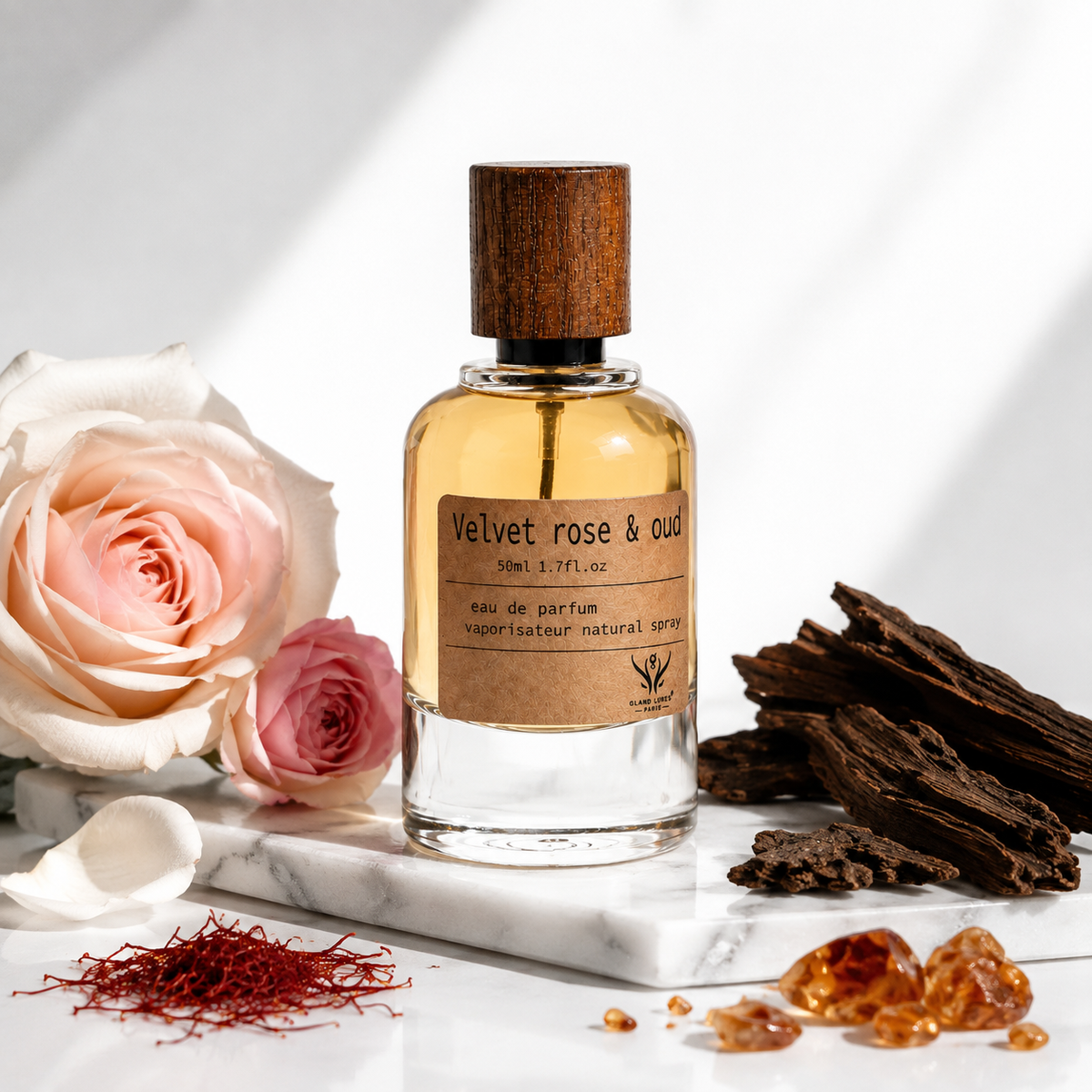 A perfume bottle labeled 'Velvet rose & oud' with amber liquid, a dark wood cap, and a kraft paper label, styled on a marble slab with a pink rose, saffron threads, and dried oud roots against a white background with soft shadows.